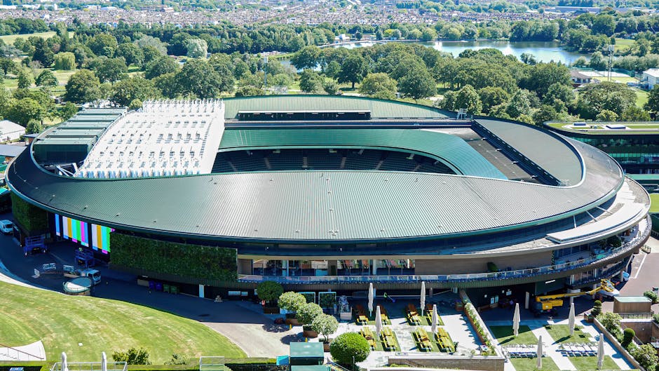 An aerial view of a large, modern sports stadium with a curved, metallic green roof covering the seating areas, surrounded by lush green trees and a landscaped garden with neatly trimmed bushes, small